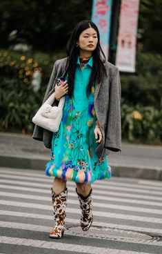 a woman in a blue dress and boots is crossing the street with her handbag