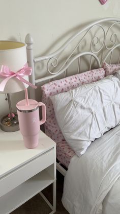 a white bed topped with pillows and a pink cup next to a night stand on top of a table