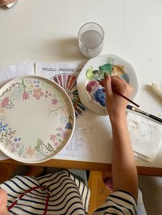 a person painting on a plate with paintbrushes and watercolors in front of them