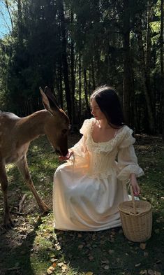 a woman in a white dress sitting on the ground next to a deer and holding a basket