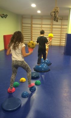 two children are playing with balls in an indoor gym area while another child watches from the sidelines