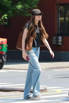 a woman walking across a cross walk wearing jeans and a brown vest with a hat on her head