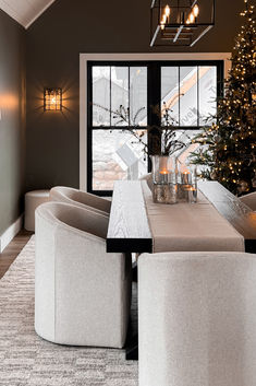 a dining room table with chairs and a christmas tree in the window behind it is lit by candles