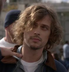 a man with long hair standing in front of another man wearing a baseball cap and jacket