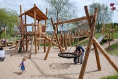 two people are standing in the sand near a wooden playground set with swings and slides