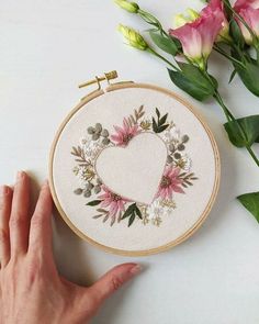 a hand is holding a cross - stitch heart on a table next to some flowers