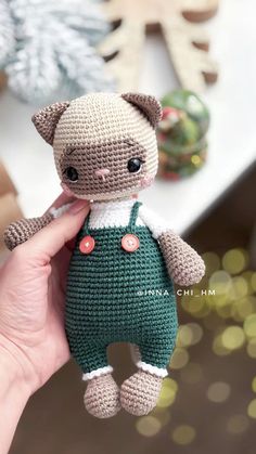 a hand holding a small crocheted cat in a green overalls and white shirt