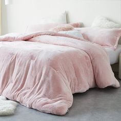 a bed with pink comforter and pillows in a white room next to a window