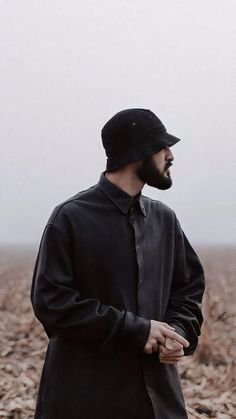 a man standing in a field wearing a black coat and hat with his hands together