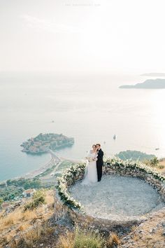 a bride and groom standing on top of a hill next to the ocean with greenery around them