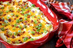 a red casserole dish with green onions, bacon and cheese in it on a wooden table