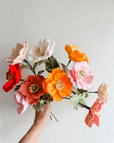 a hand holding a bunch of flowers on top of a white wall with red, orange and yellow flowers in it