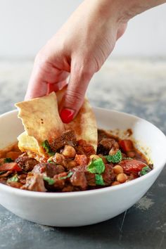 a person dipping tortilla chips into a bowl of chili with beans and meat