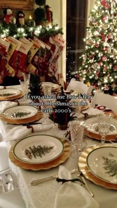 a table set for christmas dinner with plates and silverware on it, next to a decorated christmas tree