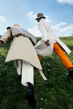 two people in white coats and orange pants are walking through the grass with their hands on each other's backs