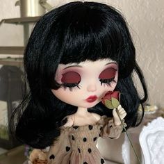 a doll with long black hair and red eyes holds a flower in her hand while standing on a table