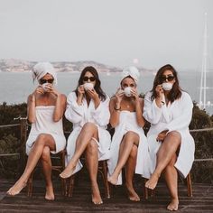 four women in white robes sitting on a wooden deck drinking coffee and looking at the camera