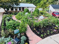 a garden with lots of plants and flowers in front of a house next to a red car