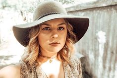 a woman wearing a hat standing in front of a wooden fence and looking at the camera