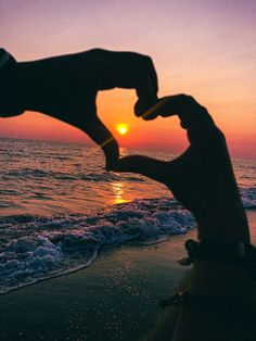 someone making a heart shape with their hands on the beach as the sun goes down