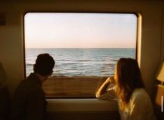 a man and woman looking out the window at the ocean from inside a train car