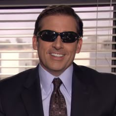 a man wearing sunglasses and a suit smiling for the camera in front of window blinds