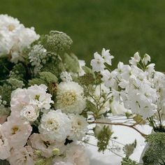 white flowers and greenery are arranged in a vase on a tablecloth covered table