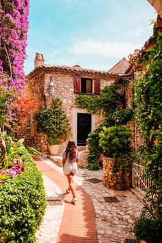 a woman walking down a brick path in front of a building with purple flowers on it