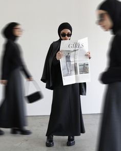 a woman in black is reading a newspaper while other people are walking past her and one person wearing a hijab