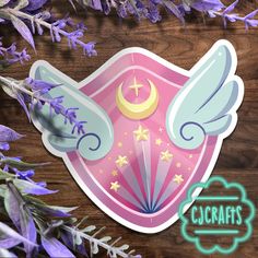 a pink sticker with angel wings and a crescent on it next to purple flowers