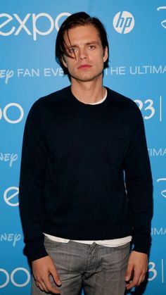 a man standing in front of a blue backdrop wearing a black sweater and grey pants