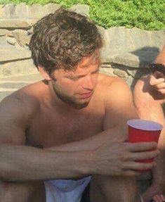 a shirtless man holding a red cup while sitting next to another man on the beach