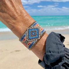 a man's hand with a beaded bracelet on the beach near the ocean