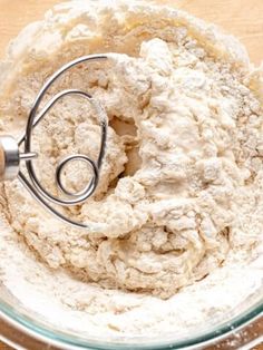 a mixing bowl filled with flour and a whisk in the middle, on top of a wooden table