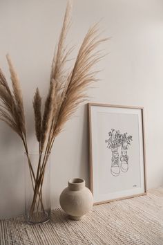 a vase with some dry grass in it next to a framed drawing on the wall