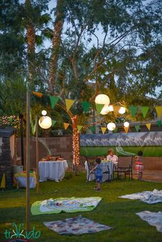 some people are sitting at a table outside under lights and flags on the grass in front of them