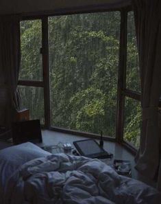 an unmade bed sitting in front of a large window with trees out the side