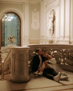 a man and woman sitting on a bench in a room with white walls, ornate columns and chandeliers