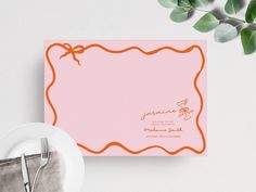 a pink card with an orange border on it next to a plate and silverware