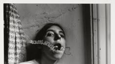 black and white photograph of woman with fake teeth in front of window, looking up at curtain