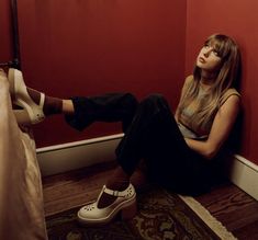 a woman sitting on the floor next to a red wall with her legs crossed and wearing white shoes