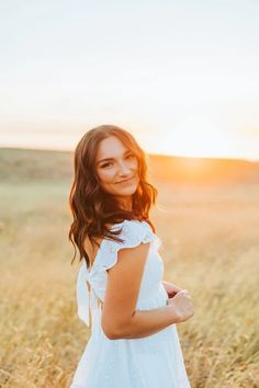 a woman in a white dress standing in tall grass with the sun setting behind her