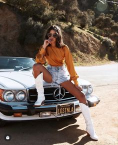 a woman sitting on the hood of a car talking on a cell phone while wearing white boots