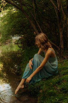 a woman sitting on the bank of a river in front of some trees and water