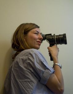 a woman holding a camera up to her face and looking into the distance with one hand