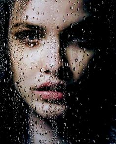 a woman looking out the window with raindrops on her face and nose, in front of a dark background