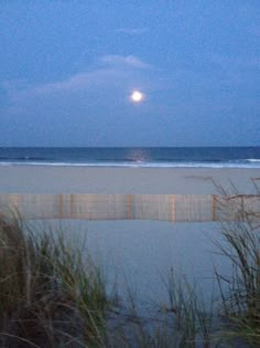 the full moon is setting over the ocean and sand dunes in front of an empty beach