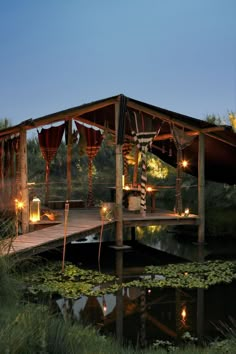 a gazebo is lit up with candles on the water and surrounded by greenery