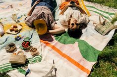 two people laying on a blanket in the grass with picnic foods and drinks around them