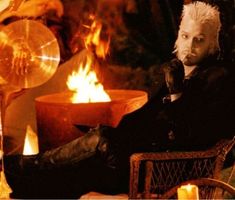 a man with white hair sitting in a chair next to a fire and some candles